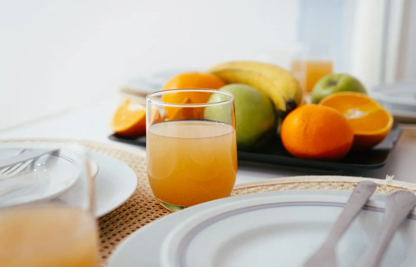 A glass of fresh juice on a breakfast table, with a tray of oranges, apples and bananas behind it