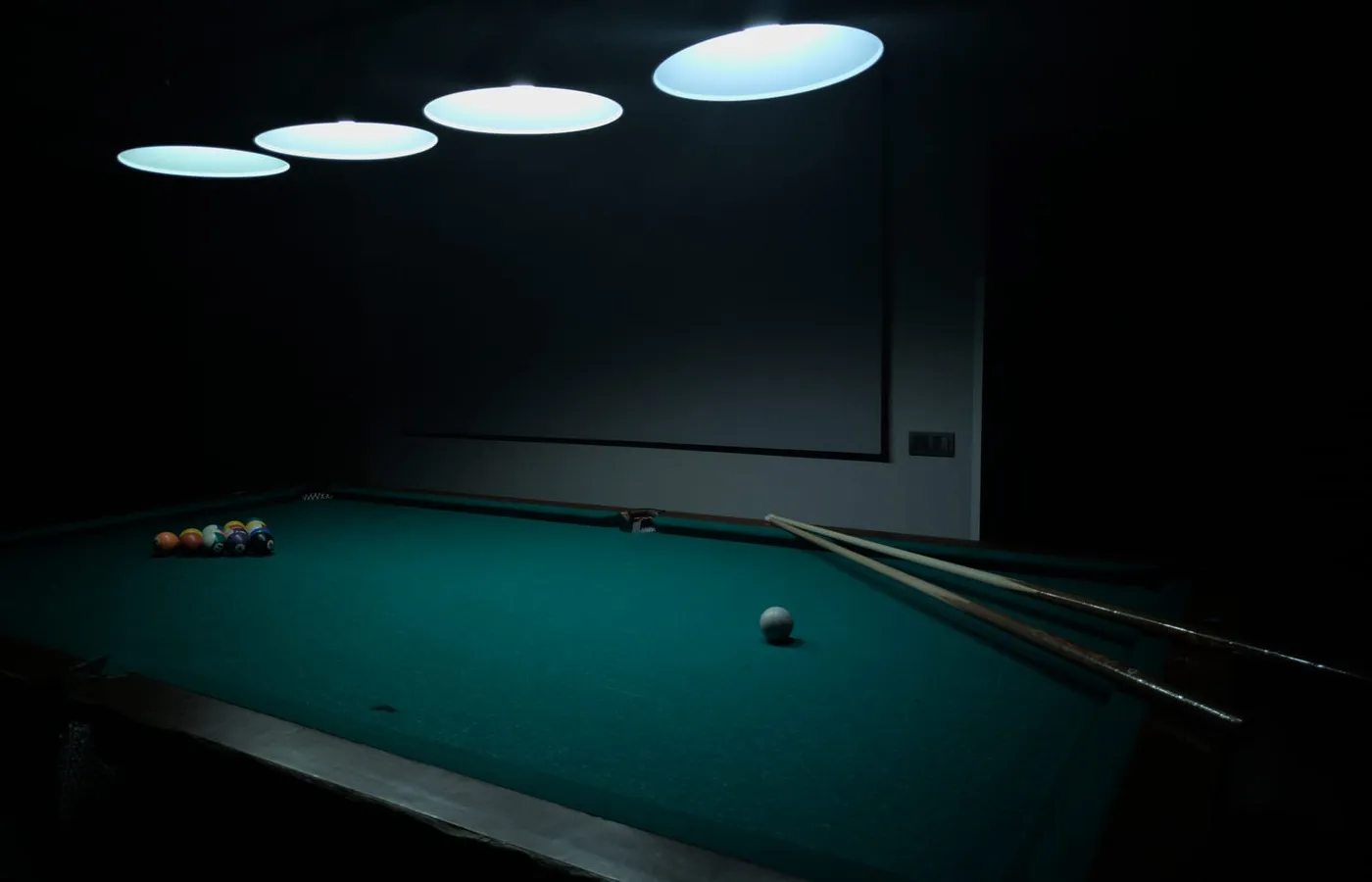 A pool table with the balls racked and two cues resting on the baize, lit by four disc pendant lights in an otherwise dark room