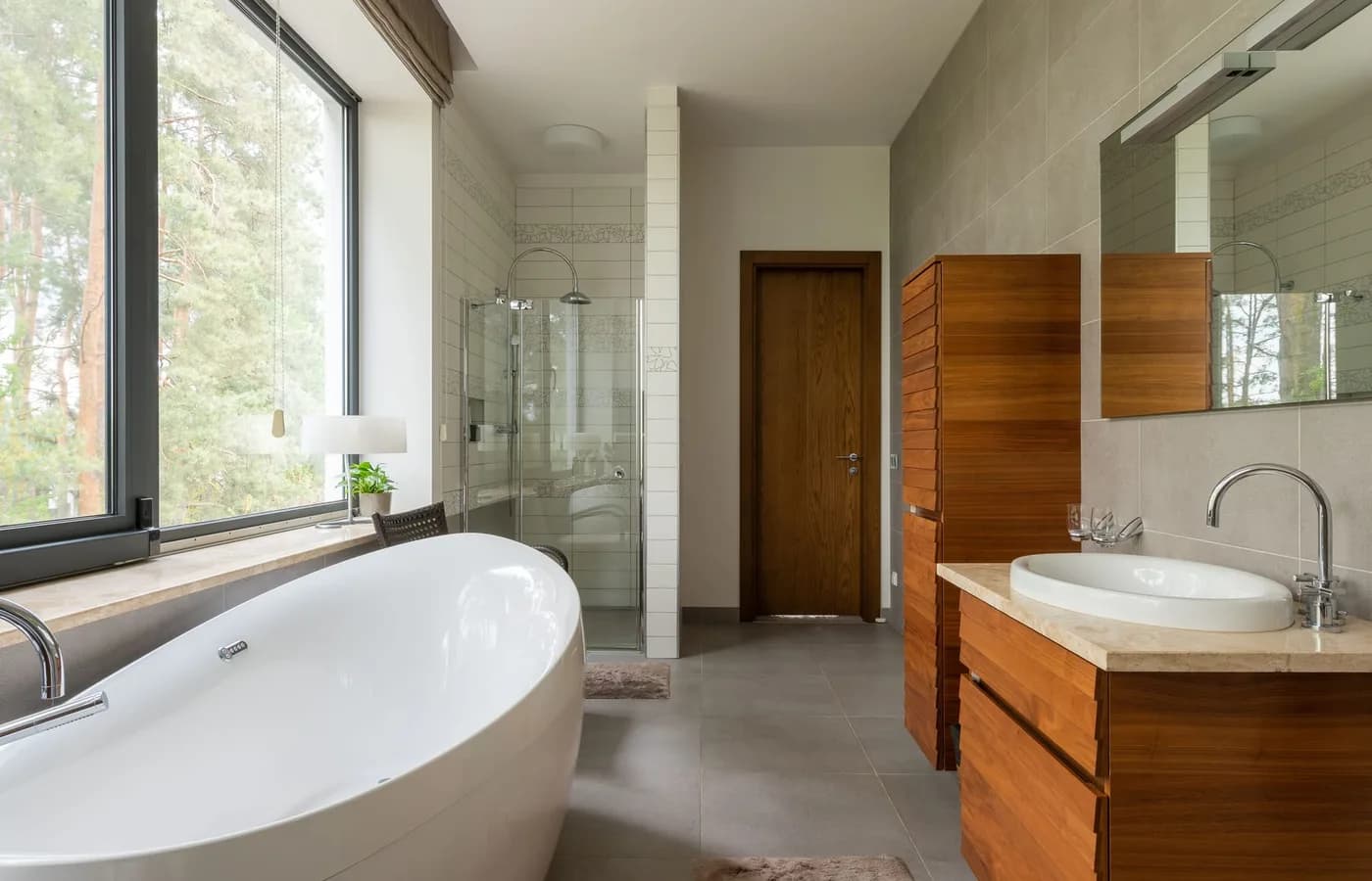 A bright modern bathroom with a freestanding white tub beside a floor-to-ceiling window, a glass shower cubicle and a wood vanity with a stone top