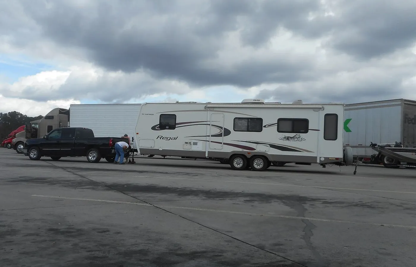 A pickup truck hitched to a travel trailer at a highway rest area, with a person working at the coupler