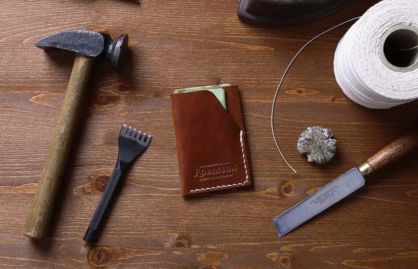 A hand-stitched leather card wallet on a dark wooden bench, surrounded by a hammer, a stitching fork, a spool of thread and an edge-bevelling knife