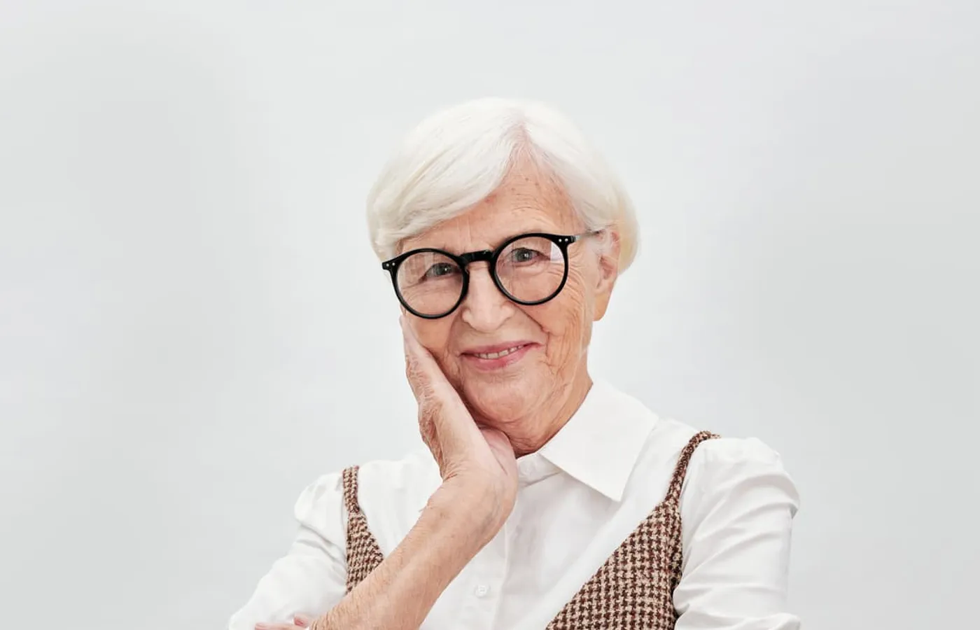 A smiling woman in her seventies with white hair and round glasses, photographed against a plain pale studio background