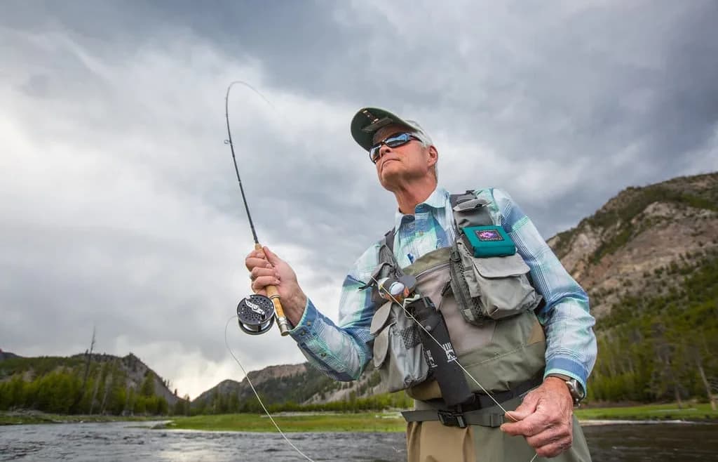 An angler in chest waders standing in a river and holding a fly rod with line in the air, under an overcast sky with wooded mountains behind