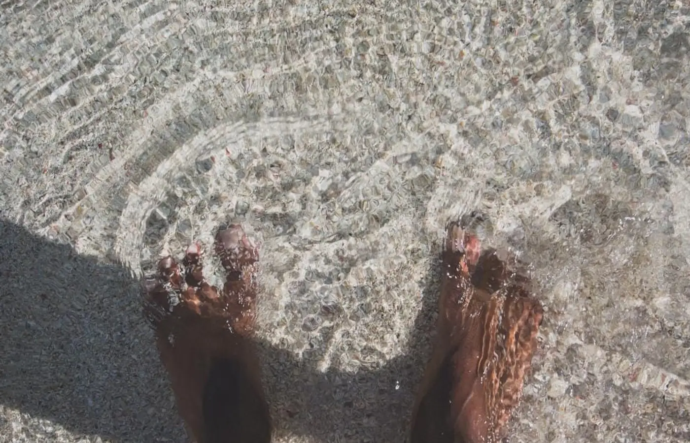 Two bare feet standing in clear shallow water over a pebbled seabed, photographed from above