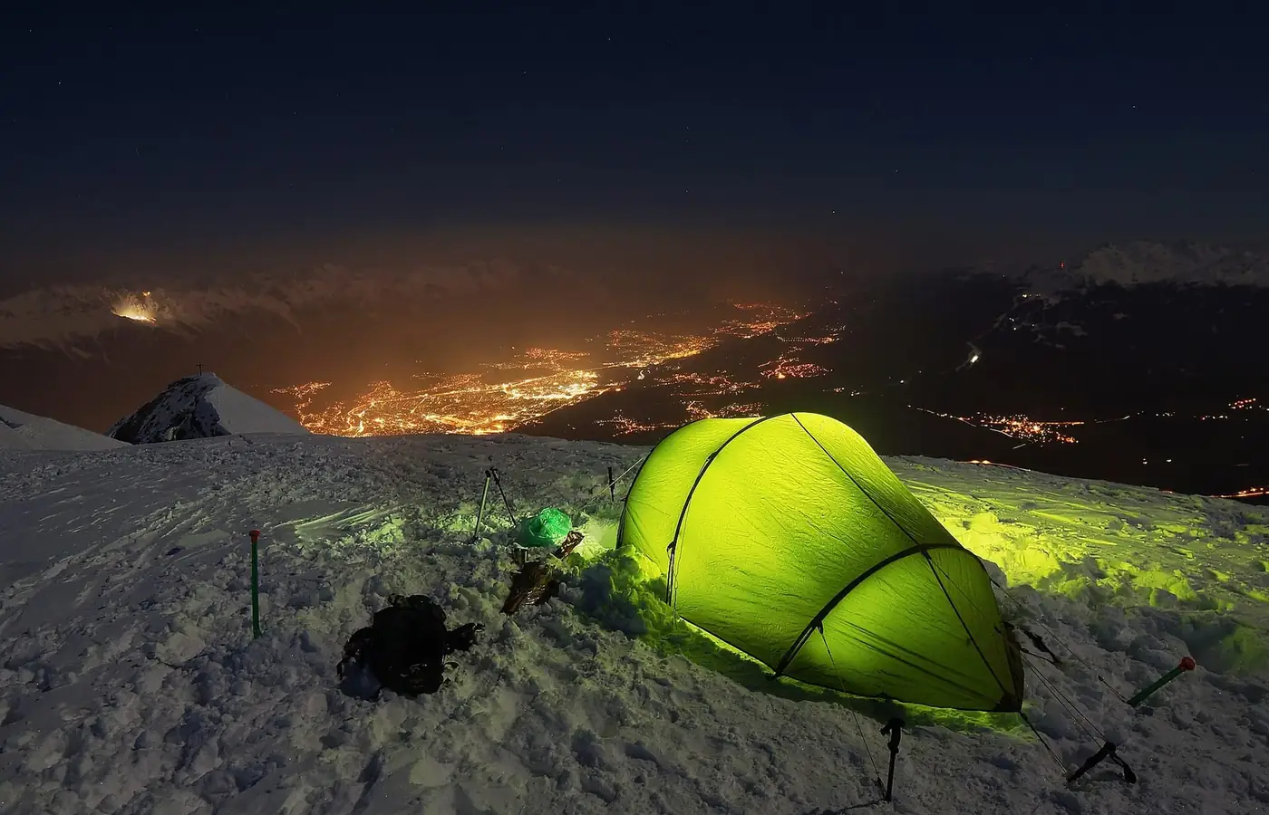 A small tent lit from inside, pitched on a snowy ridge beside a pack and trekking poles, with the lights of a valley town far below