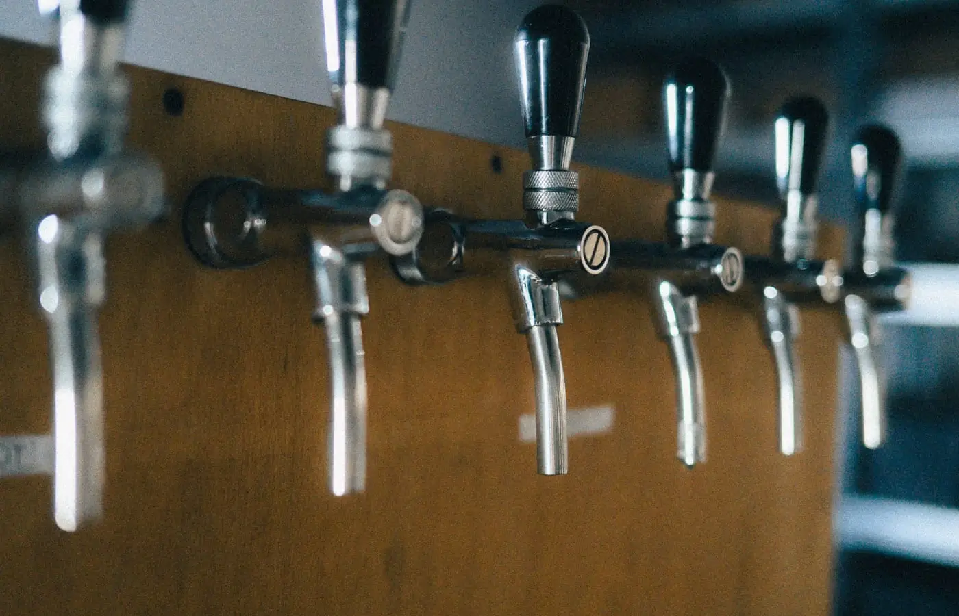 A row of chrome beer taps mounted on a wooden bar front, photographed from below against a softly blurred background